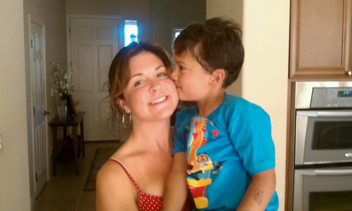 Amy, a woman in her thirties, smiles while holding a six year old child, also smiling, who is giving her a kiss on the cheek.