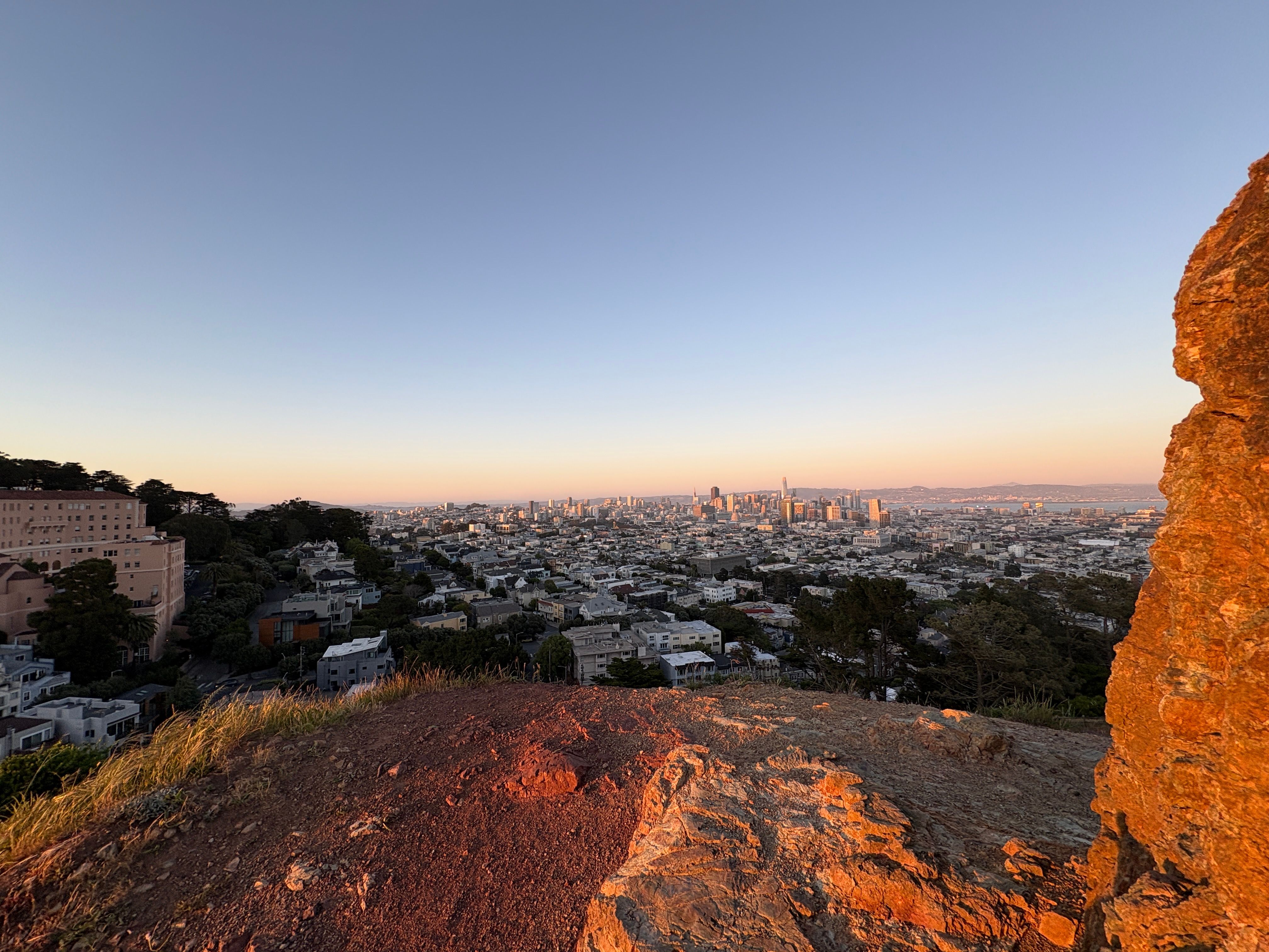 The skyline of downtown San Francisco visible, framed by a sunlit rock and the hillside. Taken from Corona Heights